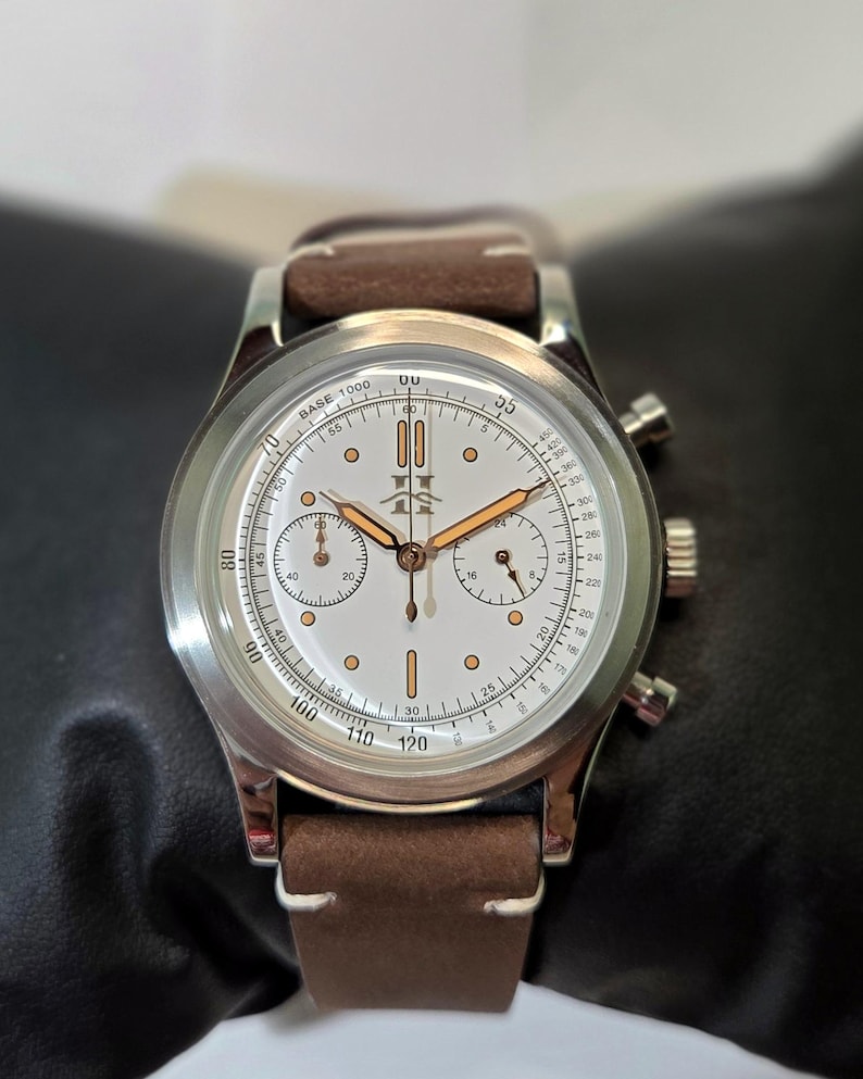 May include: A silver-tone chronograph wristwatch with a white dial, copper-colored hands, and indices. The watch features a tachymeter scale, two sub-dials, and a brown leather strap with white stitching. A logo is printed at the twelve o'clock position.