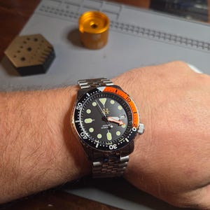May include: A stainless steel automatic diver watch with a black dial, luminous circular hour markers, and a date window at the three o'clock position. The bezel features a black and orange insert, and the watch is fitted with a metal link bracelet.
