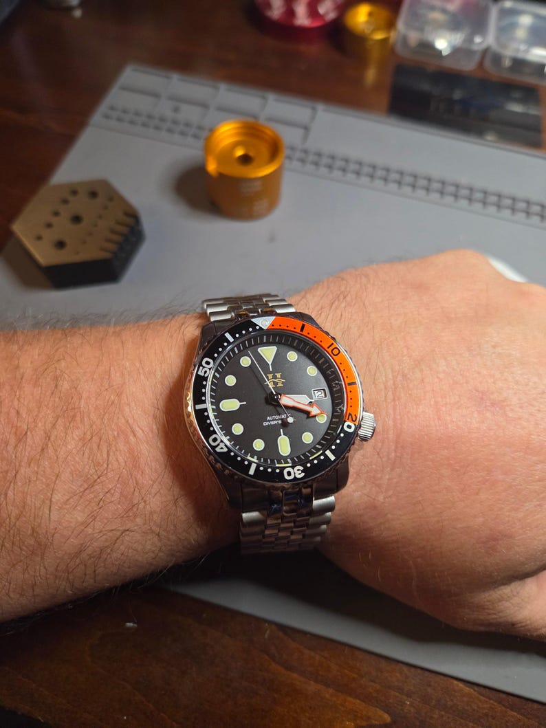 May include: A stainless steel automatic diver watch with a black dial, luminous circular hour markers, and a date window at the three o'clock position. The bezel features a black and orange insert, and the watch is fitted with a metal link bracelet.