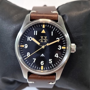 May include: A wristwatch with a round silver-tone stainless steel case, a black dial featuring white Arabic numerals, and a brown leather strap. The dial displays a gold-tone logo, a central arrow marker, and luminous hands with a thin gold-tone second hand.