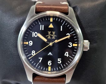 May include: A wristwatch with a round silver-tone stainless steel case, a black dial featuring white Arabic numerals, and a brown leather strap. The dial displays a gold-tone logo, a central arrow marker, and luminous hands with a thin gold-tone second hand.