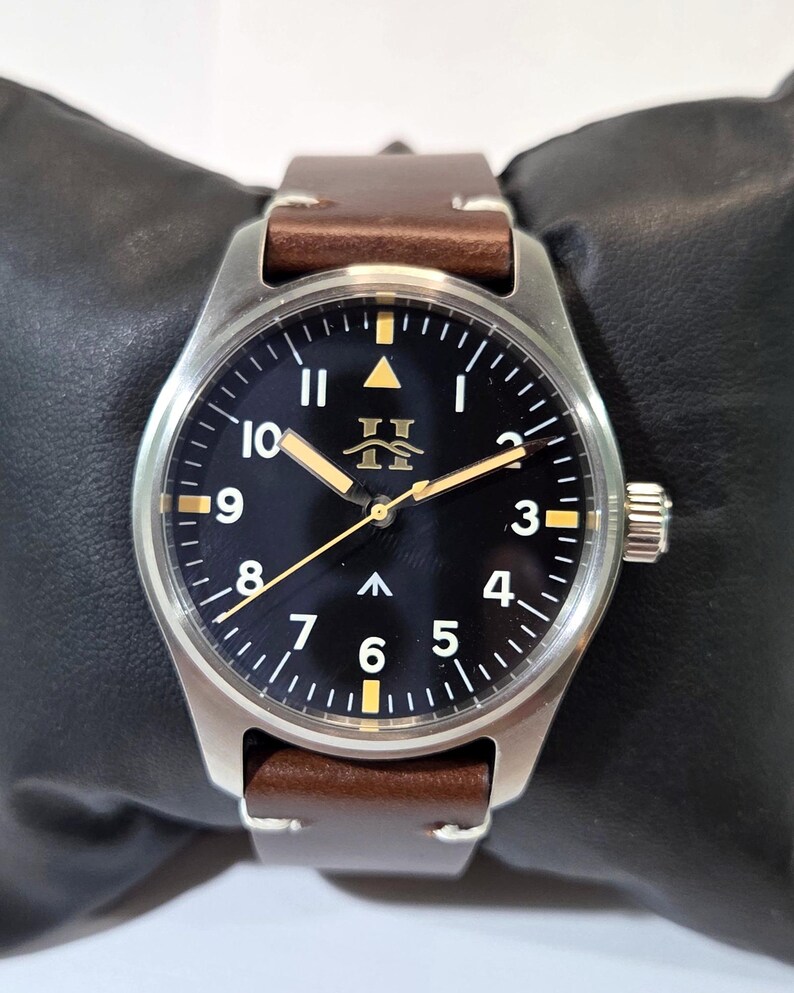 May include: A wristwatch with a round silver-tone stainless steel case, a black dial featuring white Arabic numerals, and a brown leather strap. The dial displays a gold-tone logo, a central arrow marker, and luminous hands with a thin gold-tone second hand.