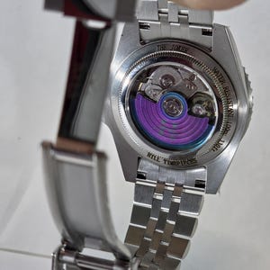 May include: A stainless steel wristwatch with an open case back revealing a purple oscillating weight. The case back is engraved with THE JOKER, WATER RESISTANT 100m 330ft, and HILL TIMEPIECES. The watch features a silver metal link bracelet.