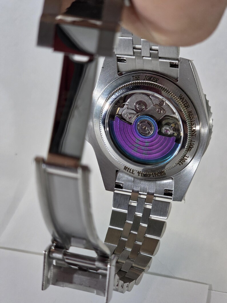 May include: A stainless steel wristwatch with an open case back revealing a purple oscillating weight. The case back is engraved with THE JOKER, WATER RESISTANT 100m 330ft, and HILL TIMEPIECES. The watch features a silver metal link bracelet.