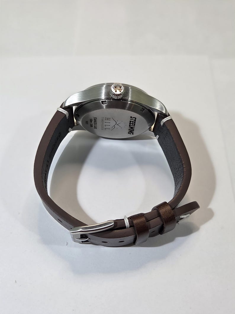 May include: A wristwatch with a brown leather strap and a silver-toned stainless steel case. The back of the watch case features the text STEELWING, HILL TIMEPIECES, STAINLESS CASE, and 50m 165ft.