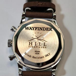 May include: A stainless steel watch case back with a brown leather strap. The back features the engraved text WAYFINDER HILL TIMEPIECES, a mountain logo, Miyota OS20, and the words Splash Resistant ONLY, with two side pushers and a crown.