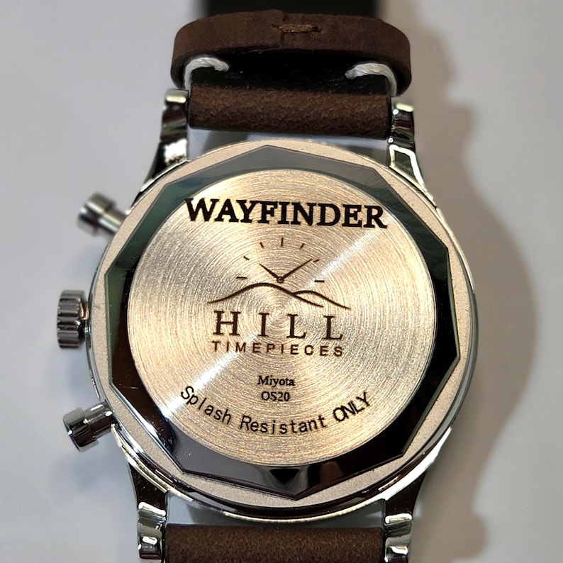 May include: A stainless steel watch case back with a brown leather strap. The back features the engraved text WAYFINDER HILL TIMEPIECES, a mountain logo, Miyota OS20, and the words Splash Resistant ONLY, with two side pushers and a crown.