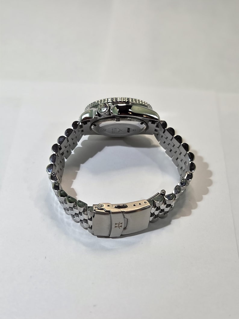 May include: A silver-toned stainless steel wristwatch featuring a multi-link jubilee-style bracelet and a fold-over deployment clasp. The watch case has a rotating bezel and a crown on the side, with a visible logo engraved on the clasp.