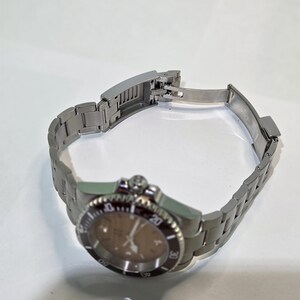 May include: A silver-toned stainless steel wristwatch featuring a brown dial and a matching metallic link bracelet. The watch includes a rotating bezel with numerical markings and a deployment clasp that is currently in an open position.