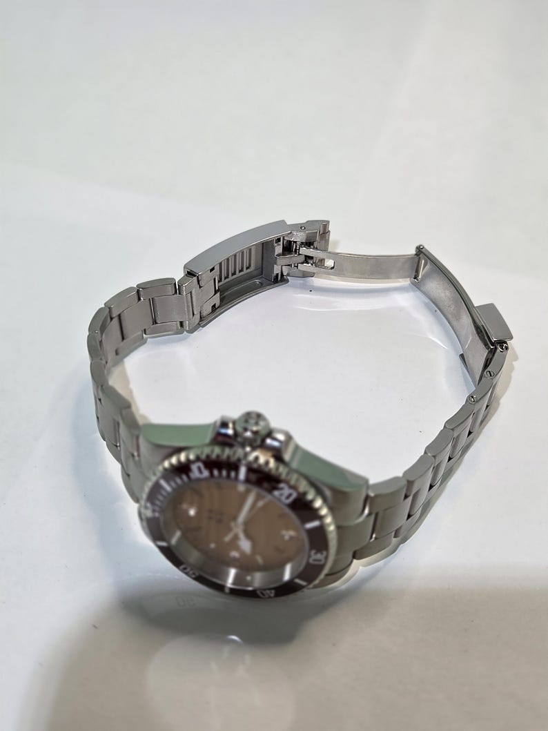 May include: A silver-toned stainless steel wristwatch featuring a brown dial and a matching metallic link bracelet. The watch includes a rotating bezel with numerical markings and a deployment clasp that is currently in an open position.