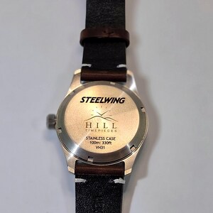 May include: A stainless steel watch case with a dark brown leather strap. The back of the case features the text STEELWING, a mountain logo, HILL TIMEPIECES, STAINLESS CASE, 100m: 330ft, and VH31. The watch has a circular silver-toned back.
