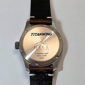 May include: A wristwatch with a dark brown leather strap and a circular titanium case back. The back features the text TITANWING, HILL TIMEPIECES, TITANIUM CASE, 100m: 330ft, and VH31, along with a graphic of a clock face above two mountain peaks.