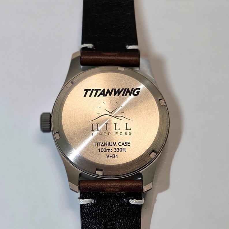 May include: A wristwatch with a dark brown leather strap and a circular titanium case back. The back features the text TITANWING, HILL TIMEPIECES, TITANIUM CASE, 100m: 330ft, and VH31, along with a graphic of a clock face above two mountain peaks.