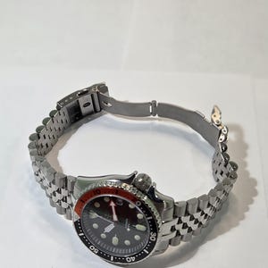 May include: A stainless steel wristwatch with a black dial, luminous hour markers, and a red and black rotating bezel. The watch features a silver-tone jubilee-style link bracelet with a fold-over clasp and a crown positioned at the four o'clock mark.