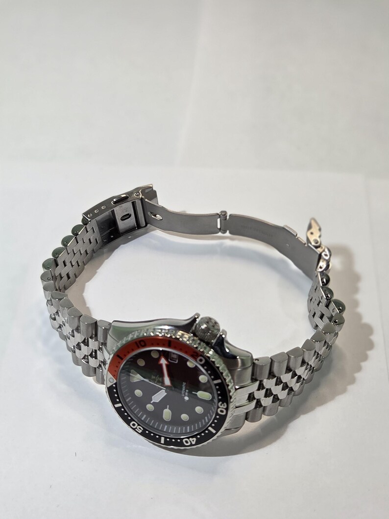 May include: A stainless steel wristwatch with a black dial, luminous hour markers, and a red and black rotating bezel. The watch features a silver-tone jubilee-style link bracelet with a fold-over clasp and a crown positioned at the four o'clock mark.