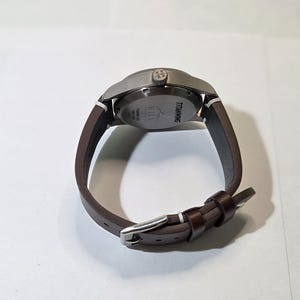 May include: A wristwatch with a dark brown leather strap and a silver-toned metal buckle. The watch case is matte gray titanium with a crown featuring a logo. The case back is engraved with the text TITANWING, HILL, and TITANIUM CASE.