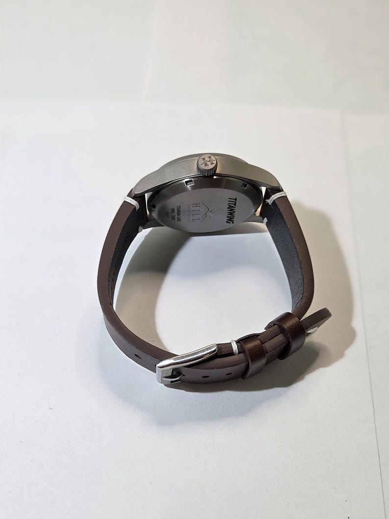 May include: A wristwatch with a dark brown leather strap and a silver-toned metal buckle. The watch case is matte gray titanium with a crown featuring a logo. The case back is engraved with the text TITANWING, HILL, and TITANIUM CASE.