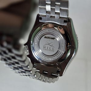 May include: A silver-toned stainless steel wristwatch with a metal link bracelet. The case back features the text BASECAMP HILL TIMEPIECES, along with engravings indicating sapphire crystal, automatic movement, and 200M water resistance.