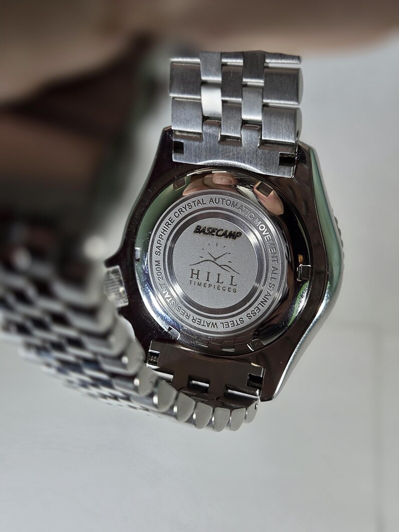 May include: A silver-toned stainless steel wristwatch with a metal link bracelet. The case back features the text BASECAMP HILL TIMEPIECES, along with engravings indicating sapphire crystal, automatic movement, and 200M water resistance.