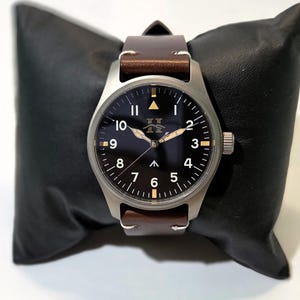 May include: A wristwatch with a matte silver-tone metal case, a black dial featuring white Arabic numerals from 1 to 10, a central upward-pointing arrow, and a logo below the 12 o'clock position. It has brown leather straps.