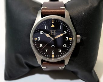 May include: A wristwatch with a matte silver-tone metal case, a black dial featuring white Arabic numerals from 1 to 10, a central upward-pointing arrow, and a logo below the 12 o'clock position. It has brown leather straps.