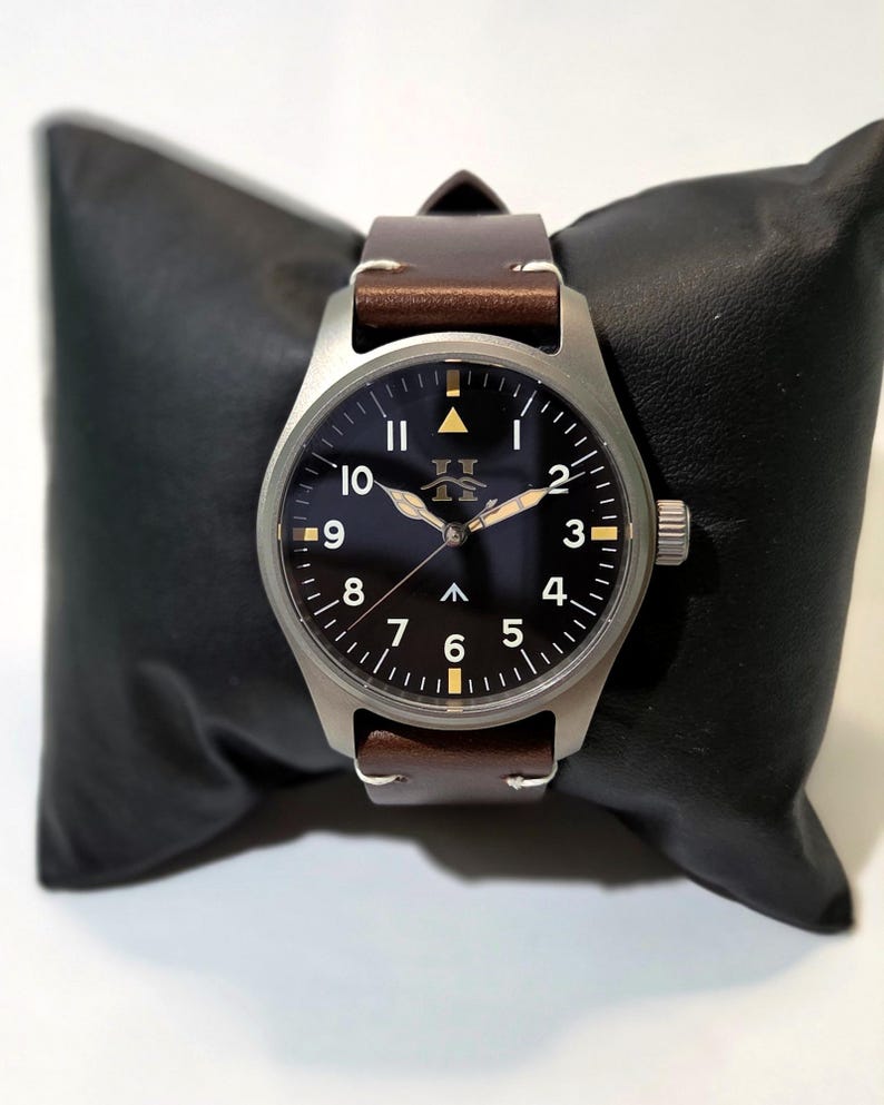 May include: A wristwatch with a matte silver-tone metal case, a black dial featuring white Arabic numerals from 1 to 10, a central upward-pointing arrow, and a logo below the 12 o'clock position. It has brown leather straps.