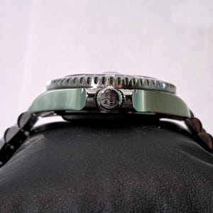 May include: A silver-toned wristwatch featuring a metallic bezel with a serrated edge and a central winding crown embossed with a logo. The watch case is polished and connects to a multi-link metal bracelet, shown from a side profile view.
