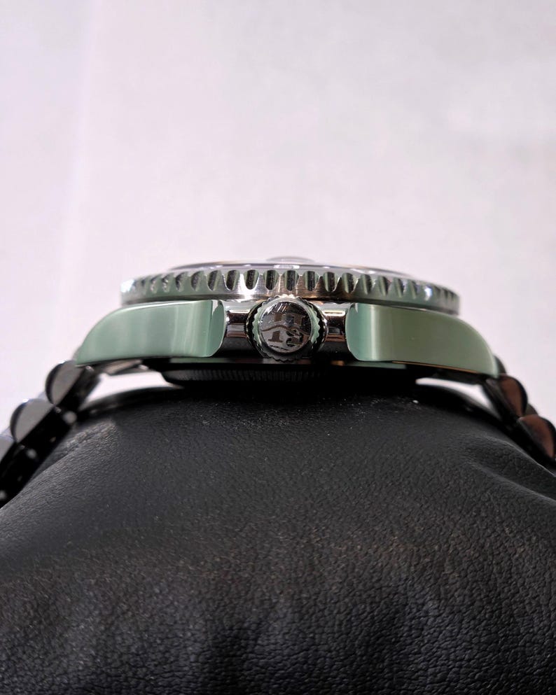 May include: A silver-toned wristwatch featuring a metallic bezel with a serrated edge and a central winding crown embossed with a logo. The watch case is polished and connects to a multi-link metal bracelet, shown from a side profile view.