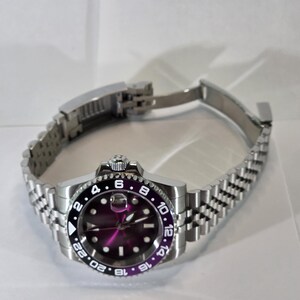 May include: A stainless steel wristwatch featuring a purple dial and a black and purple rotating bezel with 24-hour markings. The watch has a silver-tone jubilee-style link bracelet with a folding clasp and a date window with a magnifier.
