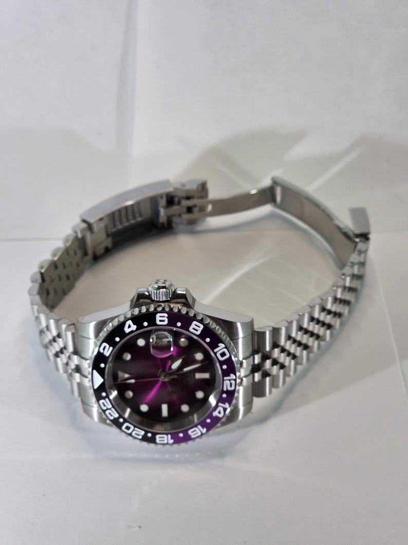 May include: A stainless steel wristwatch featuring a purple dial and a black and purple rotating bezel with 24-hour markings. The watch has a silver-tone jubilee-style link bracelet with a folding clasp and a date window with a magnifier.