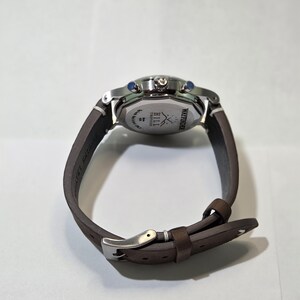 May include: A wristwatch with a brown leather strap and a silver-toned metal buckle. The back of the watch case is stainless steel, featuring the engraved text Wayfinder, Hill Timepieces, and a logo of two crossed arrows.