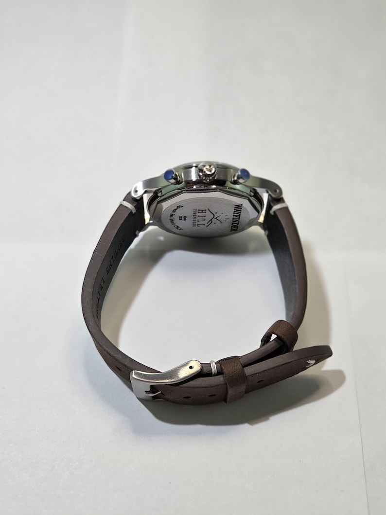 May include: A wristwatch with a brown leather strap and a silver-toned metal buckle. The back of the watch case is stainless steel, featuring the engraved text Wayfinder, Hill Timepieces, and a logo of two crossed arrows.