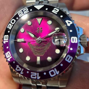 May include: A stainless steel wristwatch with a vibrant magenta sunburst dial featuring a graphic of a grinning mouth. The bezel is split between black and purple with white numerals. It includes luminous hour markers, silver hands, and a date window.