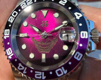 May include: A stainless steel wristwatch with a vibrant magenta sunburst dial featuring a graphic of a grinning mouth. The bezel is split between black and purple with white numerals. It includes luminous hour markers, silver hands, and a date window.