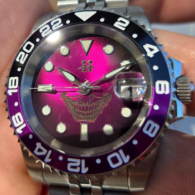May include: A stainless steel wristwatch with a vibrant magenta sunburst dial featuring a graphic of a grinning mouth. The bezel is split between black and purple with white numerals. It includes luminous hour markers, silver hands, and a date window.