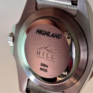 May include: The stainless steel case back of a wristwatch features a circular brushed metal center with the text HIGHLAND HILL TIMEPIECES, a mountain graphic with clock hands, 200m, and NH35 engraved in black lettering.