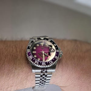 May include: A stainless steel wristwatch featuring a deep purple dial, a silver-tone jubilee bracelet, and a rotating bezel with a black and purple insert marked with 24-hour numerals. The watch includes a date window with a magnifying lens.