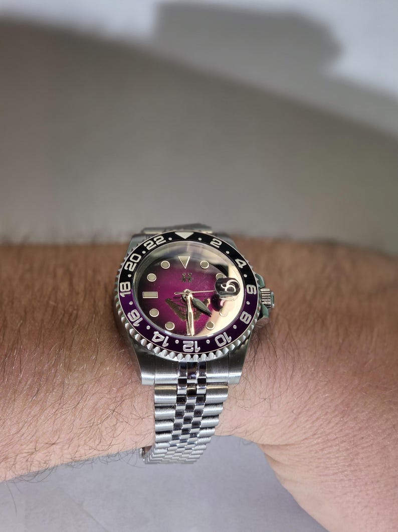 May include: A stainless steel wristwatch featuring a deep purple dial, a silver-tone jubilee bracelet, and a rotating bezel with a black and purple insert marked with 24-hour numerals. The watch includes a date window with a magnifying lens.