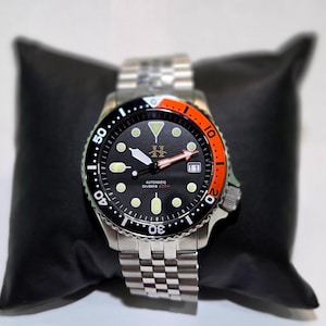 May include: An automatic diver's watch with a black dial, luminous circular indices, and a date window at the three o'clock position. The bezel features a black and orange insert with white numerals. It has a stainless steel link bracelet.