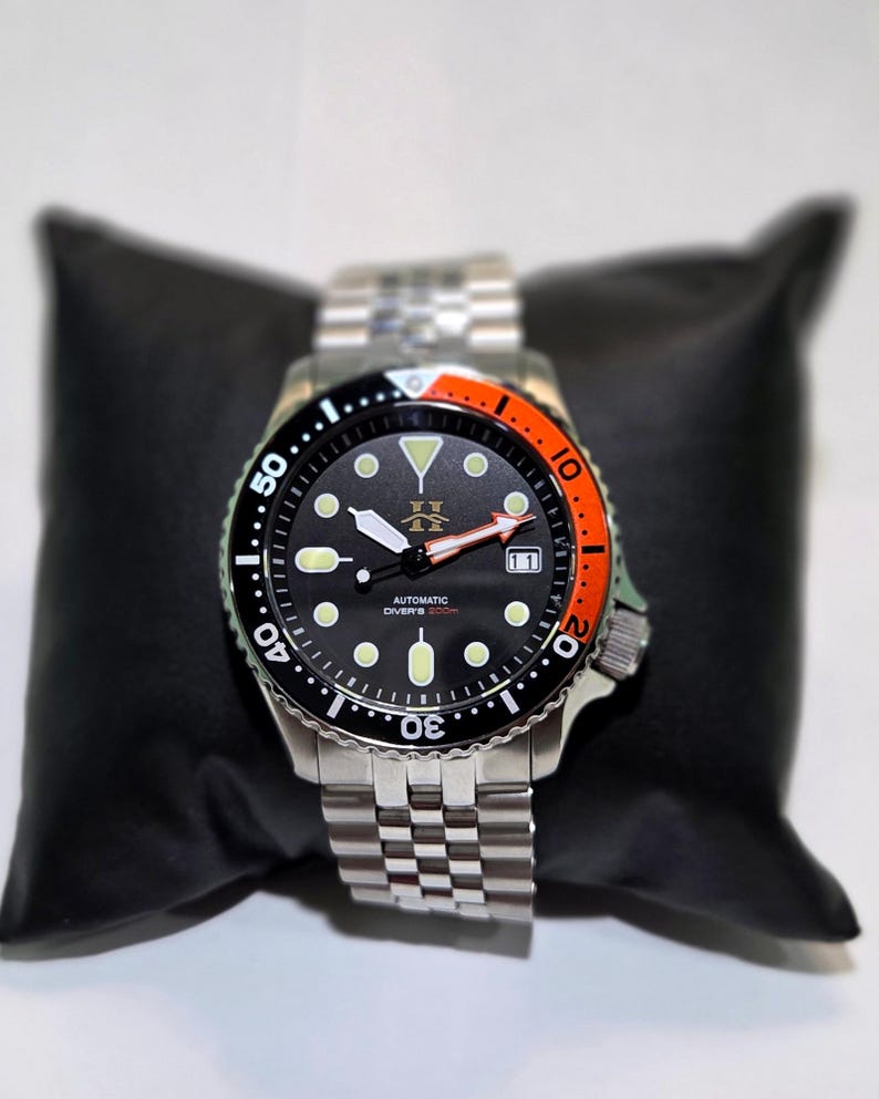 May include: An automatic diver's watch with a black dial, luminous circular indices, and a date window at the three o'clock position. The bezel features a black and orange insert with white numerals. It has a stainless steel link bracelet.