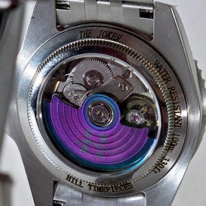 May include: A silver-toned watch case back with an exhibition window revealing a mechanical movement featuring a vibrant purple and blue iridescent rotor. The metal casing is engraved with THE JOKER, WATER RESISTANT 100m 330ft, and HILL TIMEPIECES.