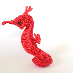 Könnte beinhalten: Ein gehäkeltes rotes Seepferdchen mit einem eingerollten Schwanz und einem kleinen schwarzen Auge. Das Seepferdchen ist aus Garn gefertigt und ein handgemachtes Spielzeug.