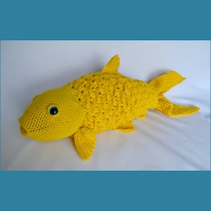Puede incluir: Un juguete de pescado amarillo tejido a crochet con ojos azules. El pez está hecho con un punto texturizado y tiene una cola larga.