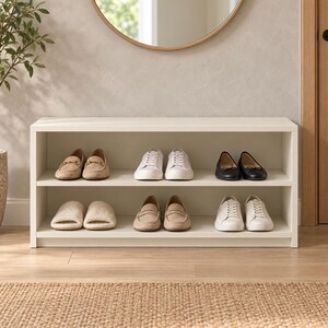 Può includere: Una scarpiera bianca a due livelli con design aperto. Il ripiano superiore contiene un paio di mocassini marrone chiaro, scarpe da ginnastica bianche e ballerine nere. Il ripiano inferiore contiene un paio di pantofole color crema.