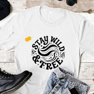 May include: A white short-sleeved crew neck t-shirt featuring a circular black graphic design in the centre. The graphic contains the text "STAY WILD & FREE" written in a bold, stylised font, surrounded by swirling lines and small star accents.