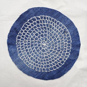 Könnte beinhalten: Ein kreisförmiger Denim-Patch mit einem weißen, gestickten Spitzendesign. Der Denim hat eine mittelblaue Farbe, und die Stickerei ist strahlend weiß. Das Design ist auf dem Patch zentriert und hat ein detailliertes, spitzenartiges Aussehen.