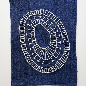 May include: A rectangular piece of dark blue denim fabric featuring a hand-embroidered white thread design. The embroidery consists of two concentric oval shapes connected by short, evenly spaced vertical stitches, creating a radial pattern.