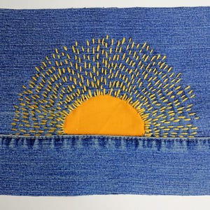 May include: A rectangular piece of blue denim fabric featuring a semi-circular orange fabric patch at the bottom center. Yellow embroidery thread creates radiating sun rays extending upward from the orange patch across the denim surface.