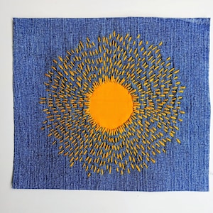 Op de afbeelding: Een rechthoekig stuk blauwe spijkerstof met een centrale ronde oranje stoffen patch. De patch is omringd door uitstralende gele borduursteken die zich in een zonnestraalpatroon over het oppervlak van de spijkerstof uitstrekken.
