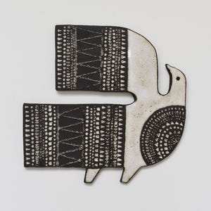 May include: A flat ceramic wall plaque shaped like a stylized bird. The piece features a cream-colored head and body with a black wing and tail section. The black areas are decorated with intricate white geometric patterns, including triangles and dots.